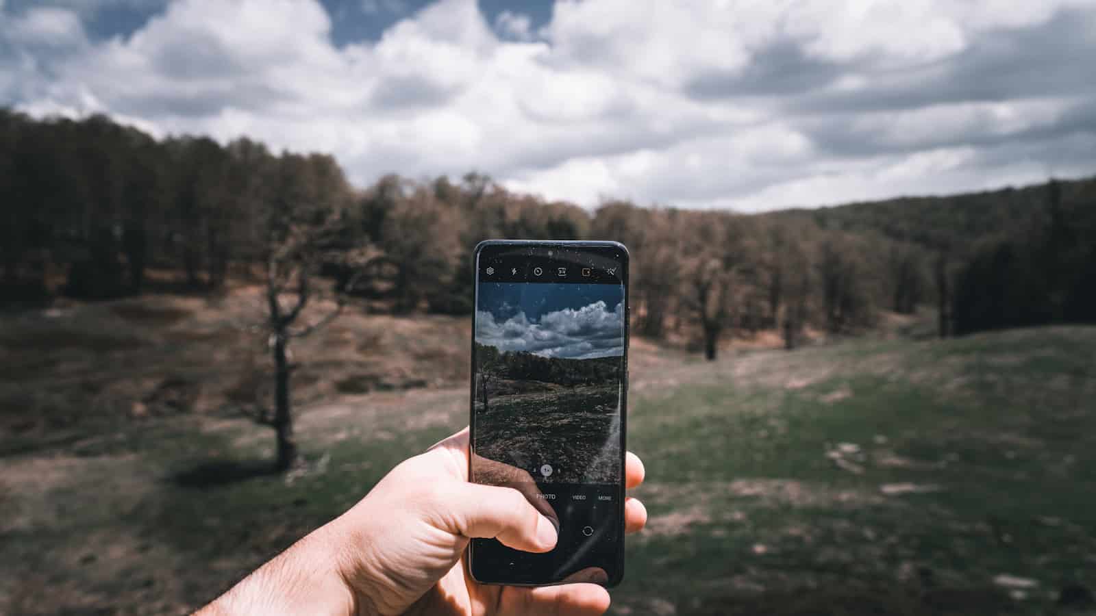 A hand holding up an Android phone to photograph an open field under a cloudy sky.