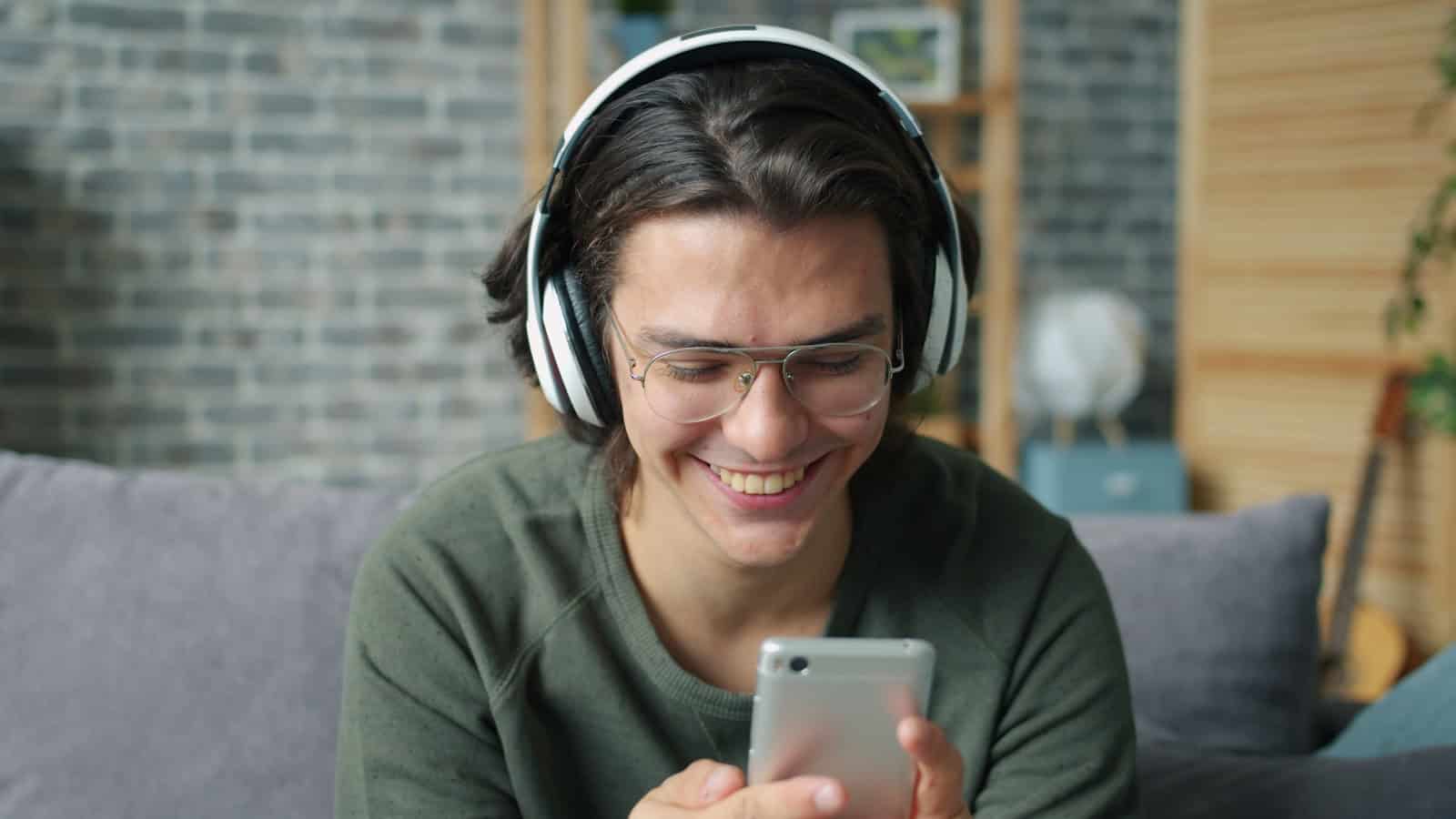 A man in glasses and over-ear headphones smiling at the phone in his hand.