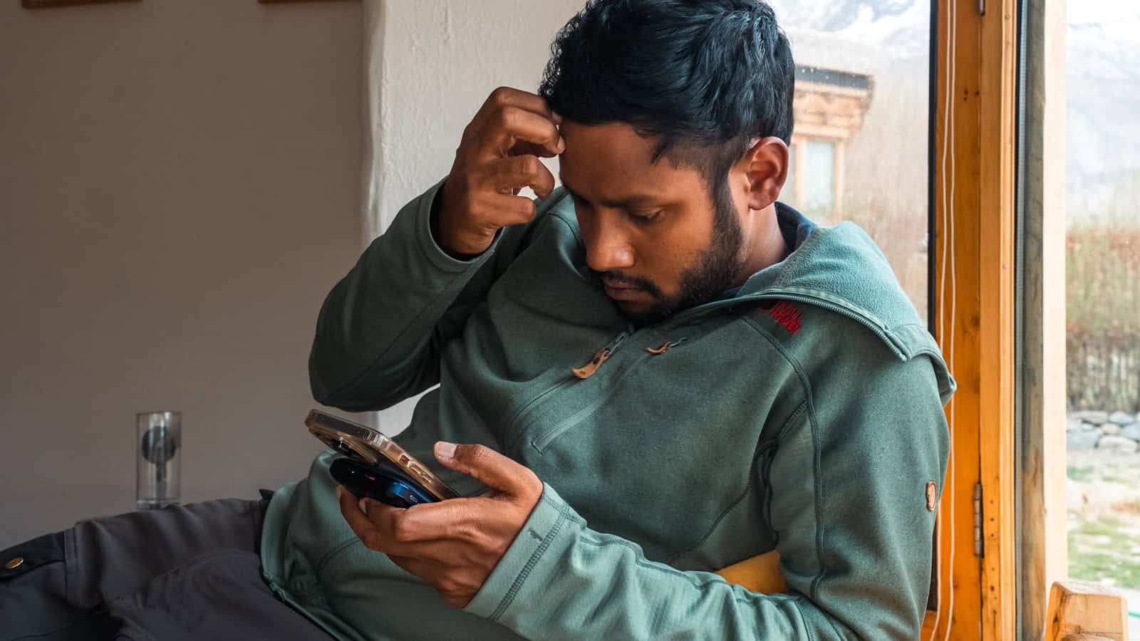 A man frowning at the phone in his hand while standing beside a window.