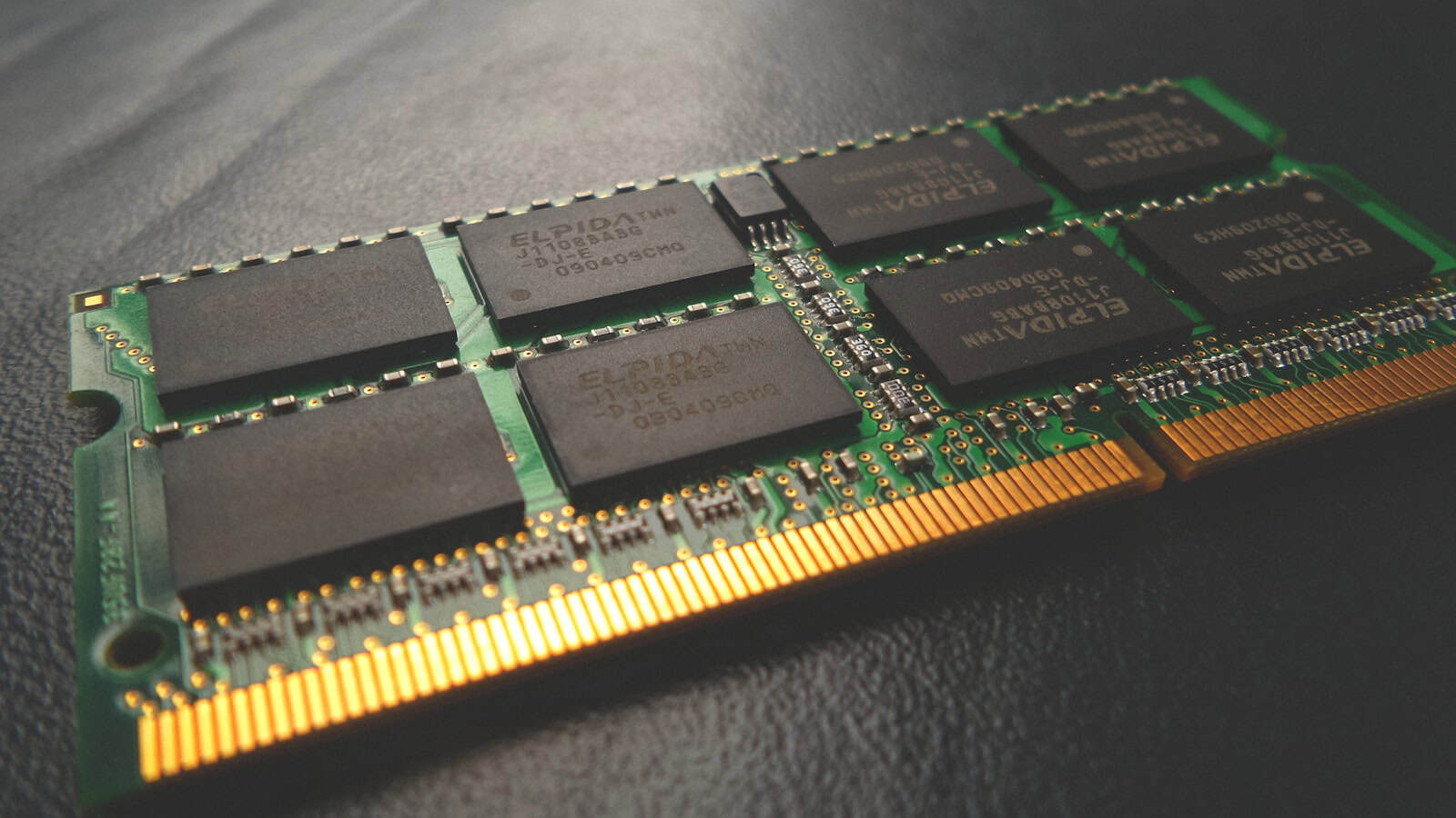 A single laptop memory module lying at an angle on a dark surface, its chips and gold contacts visible.