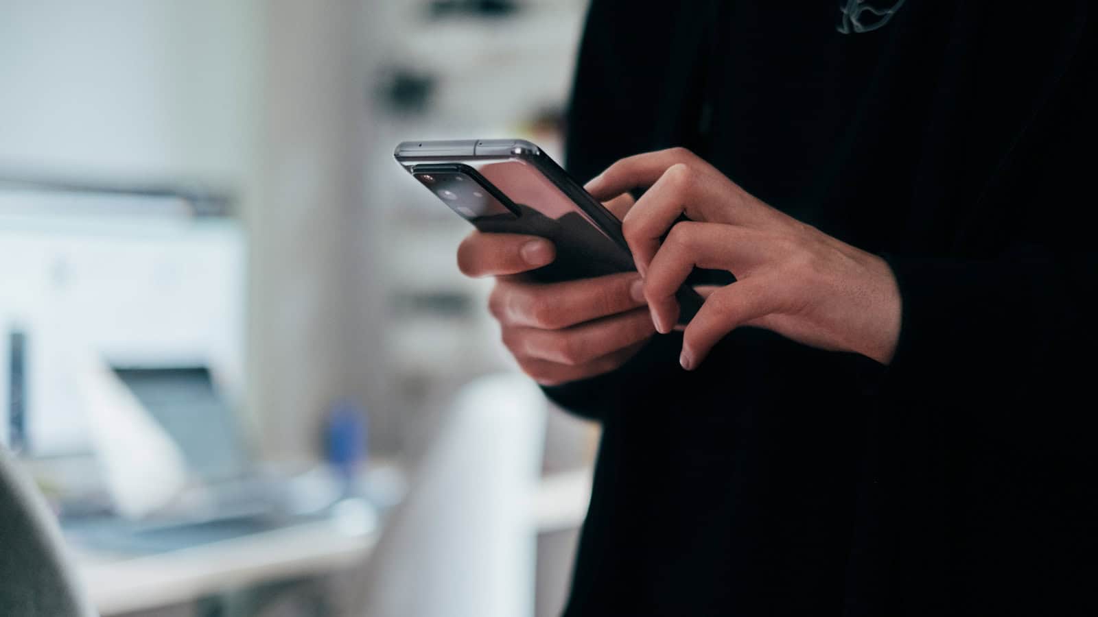 Two hands holding a dark phone and tapping the screen in an office.