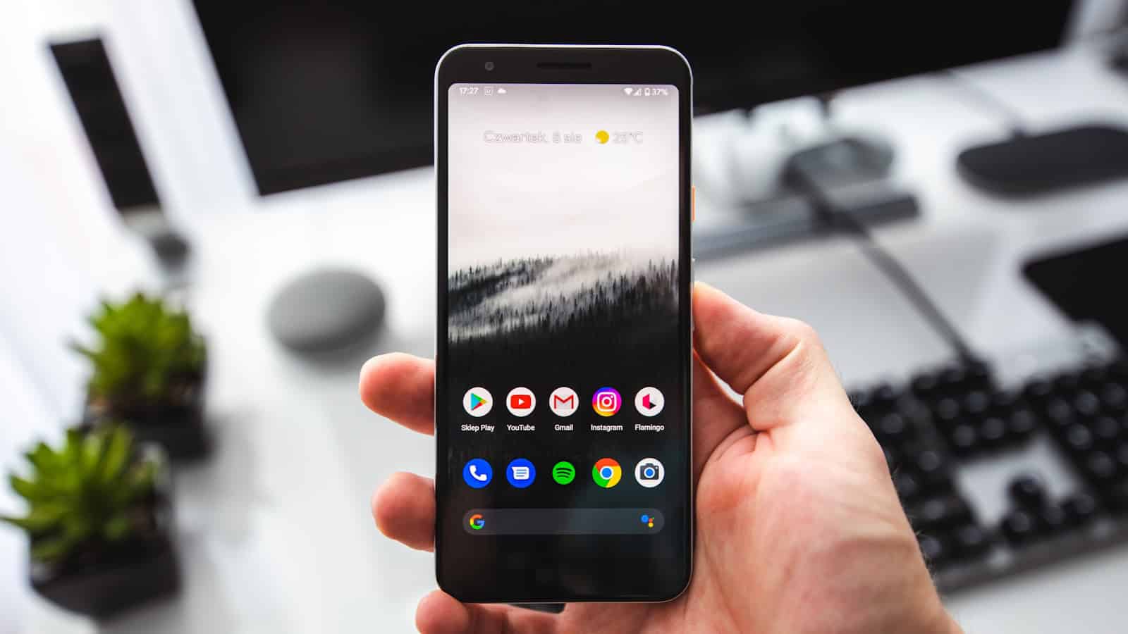 A hand holding an Android phone showing its home screen of Google app icons beside a laptop keyboard.