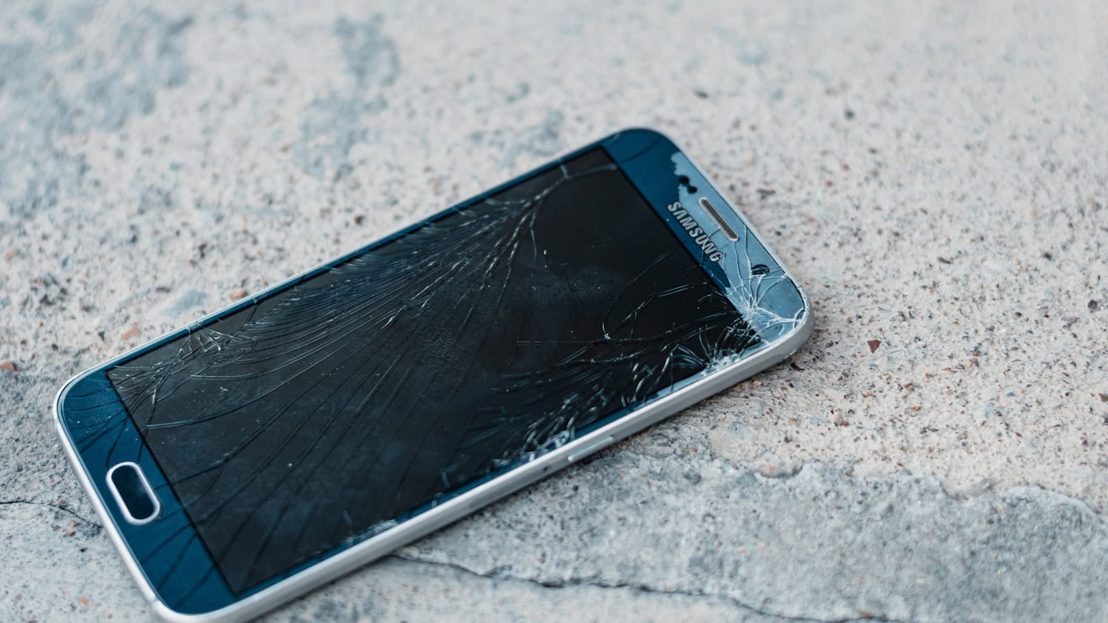 A phone lying face up on concrete with its screen shattered across the whole panel.