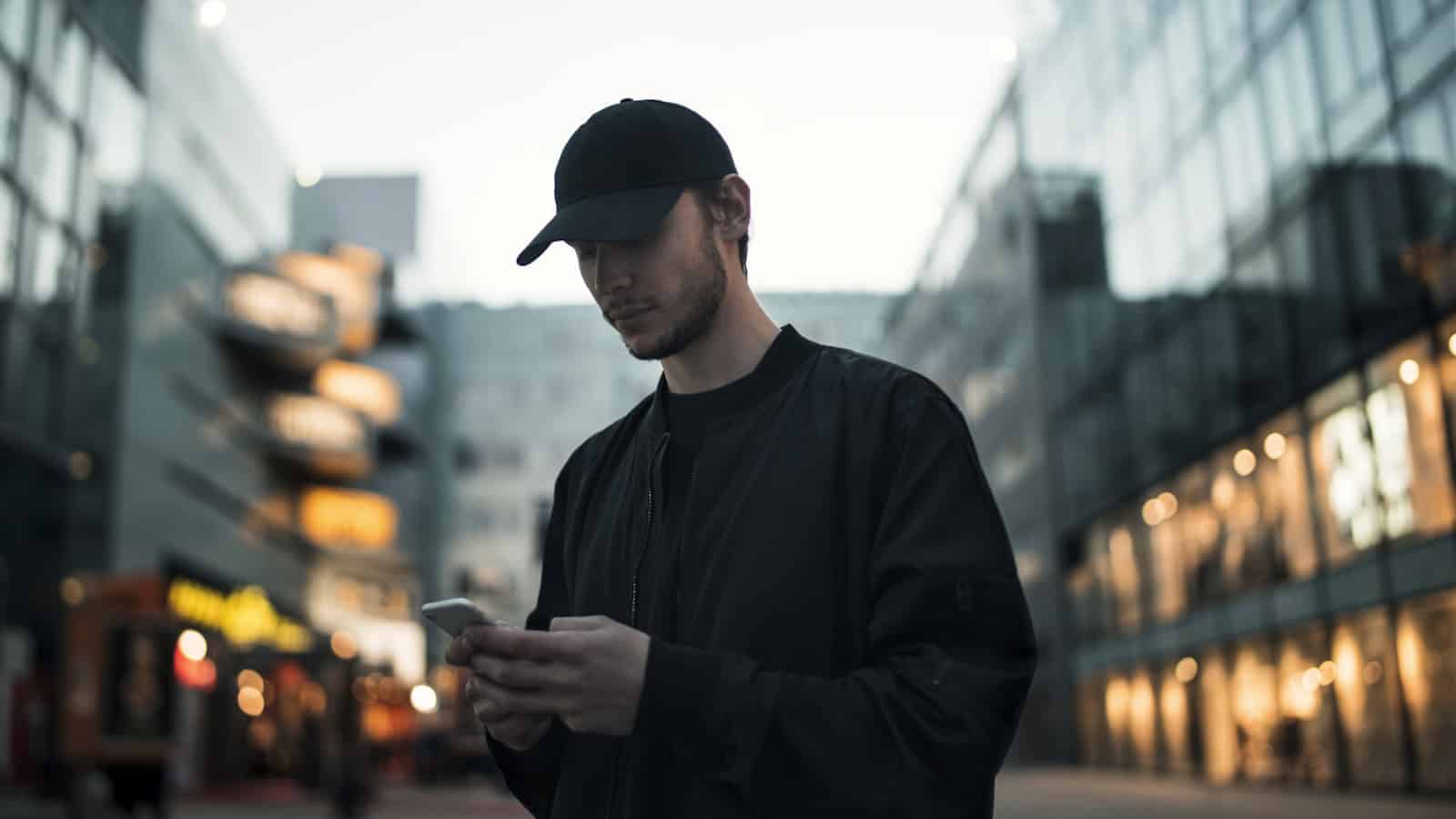 A man in a cap using his phone on a city street after dark.
