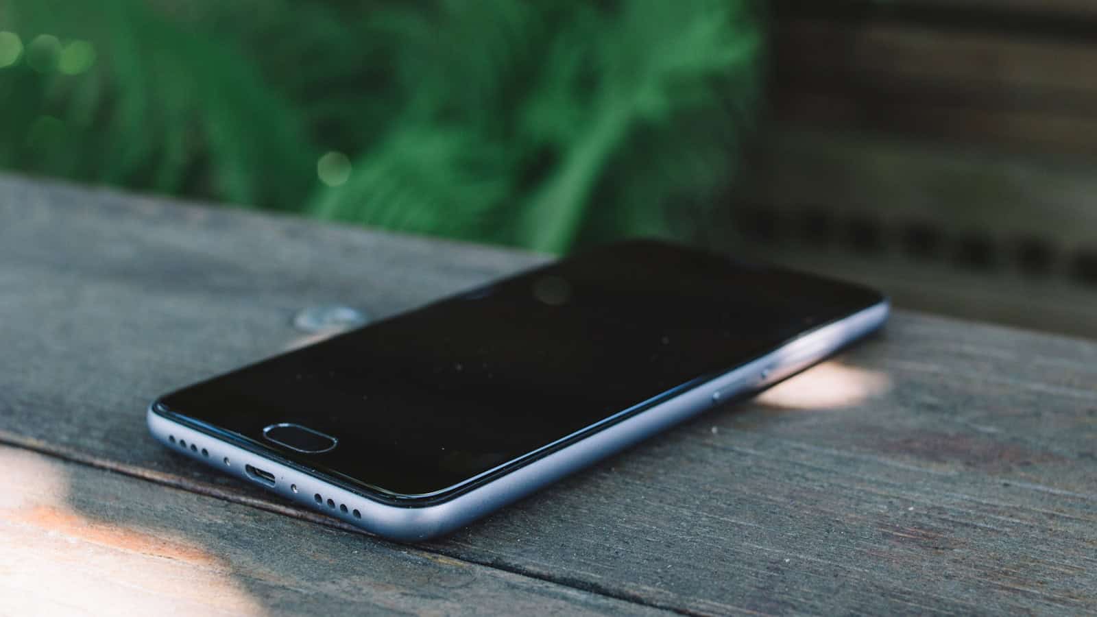 A dark phone lying face down on a weathered wooden table with greenery behind it.