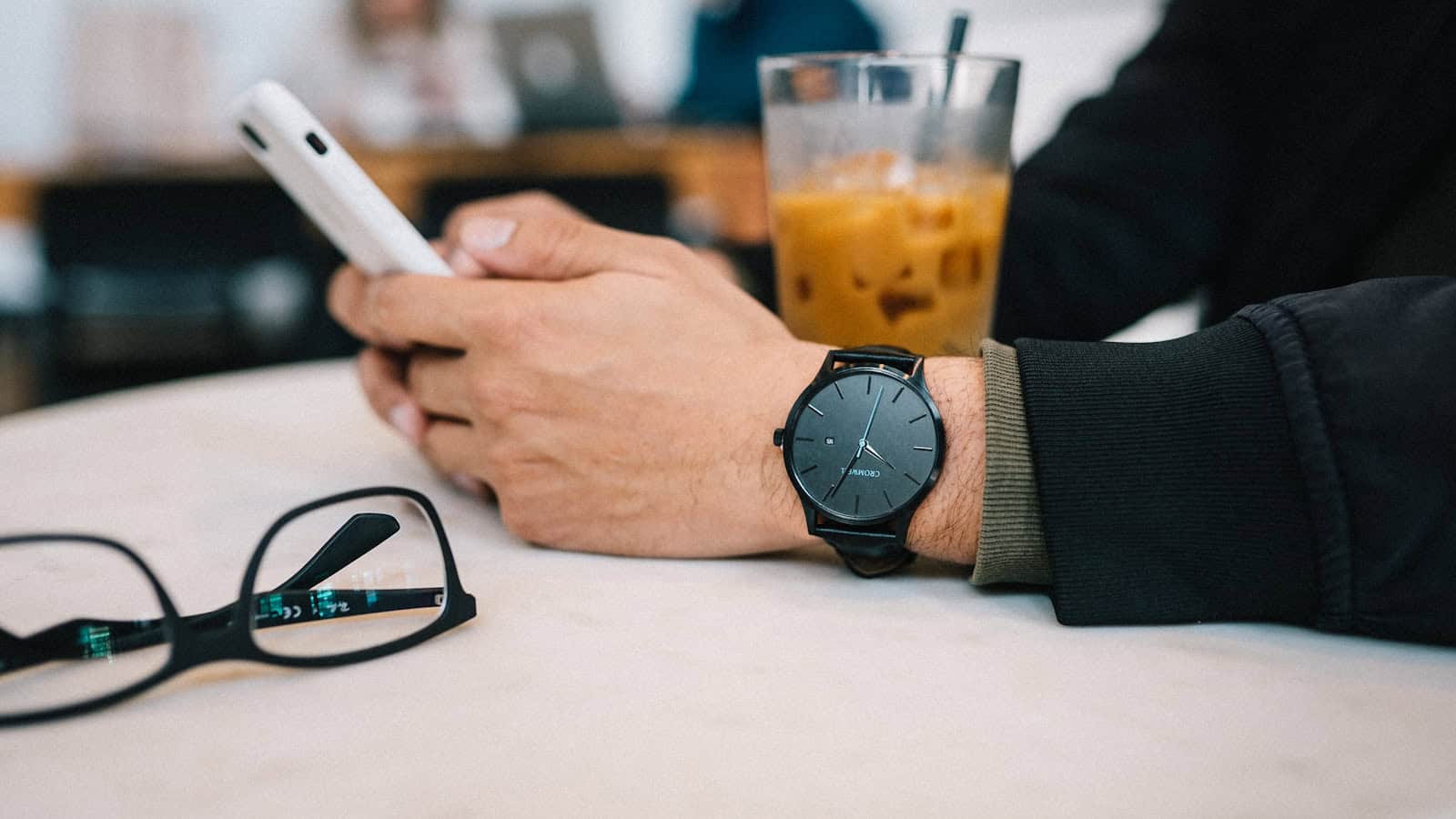 A man using his phone at a cafe table beside an iced coffee and a pair of glasses.