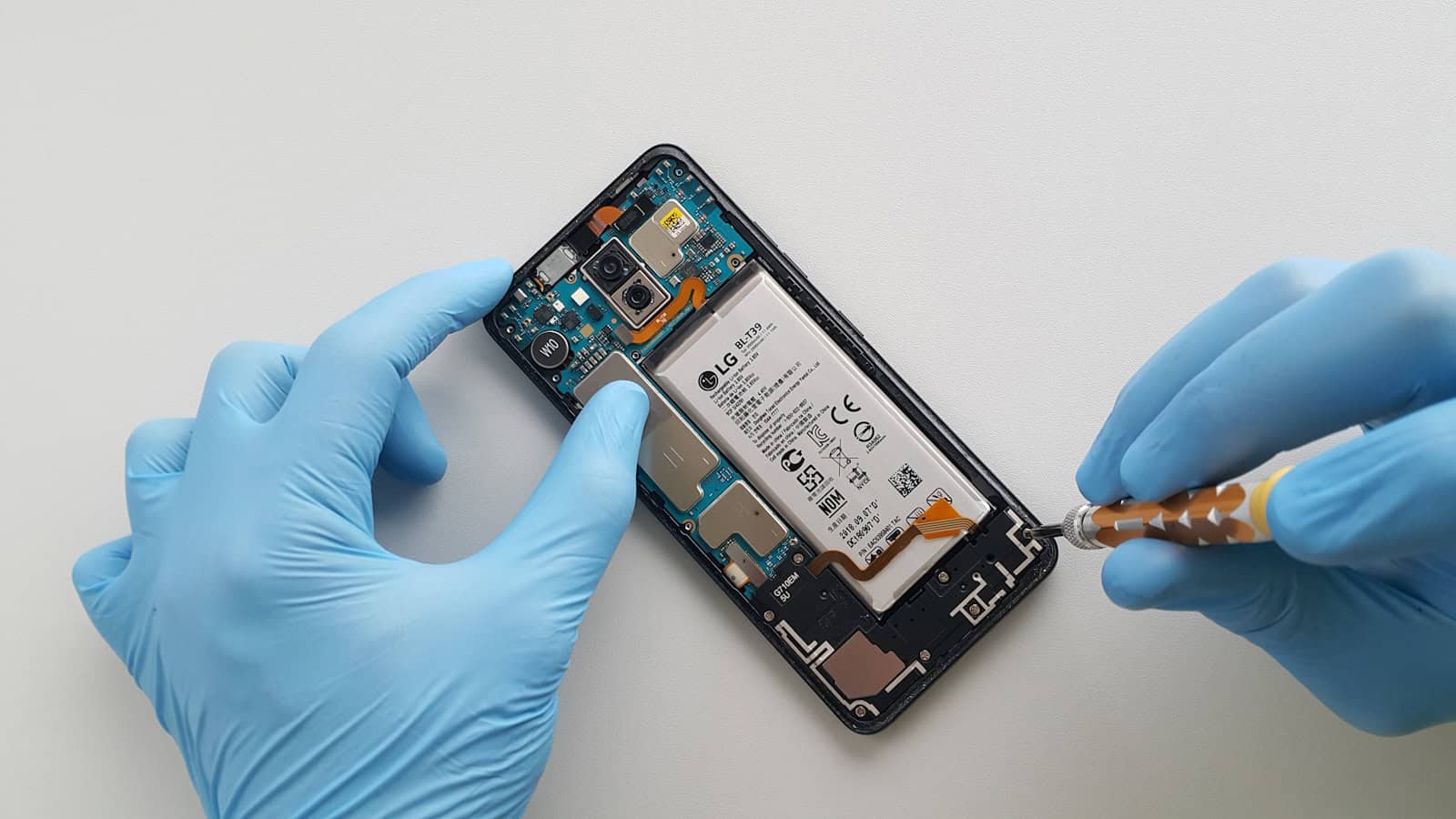 Gloved hands using a screwdriver inside an opened Android phone, the battery visible.