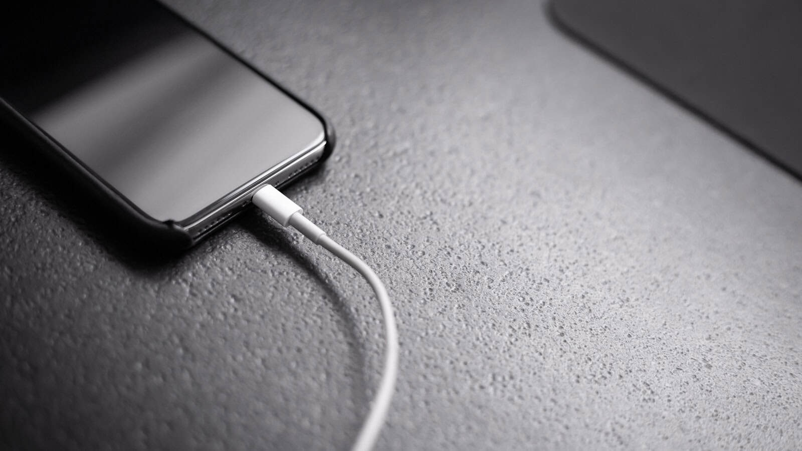 A black phone face down on a dark textured surface with a white charging cable plugged into its port.