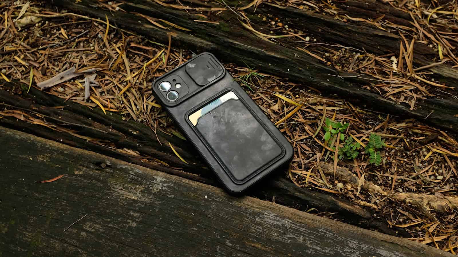 A phone in a black wallet case lying face down on bare ground beside a wooden edge.