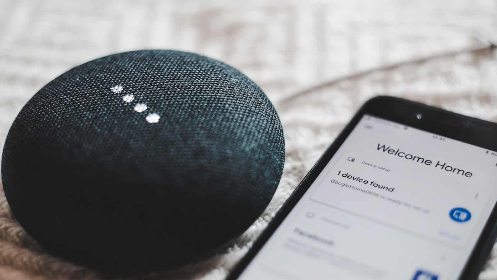 A phone showing a device-discovery screen lying beside a dark fabric-covered smart speaker.