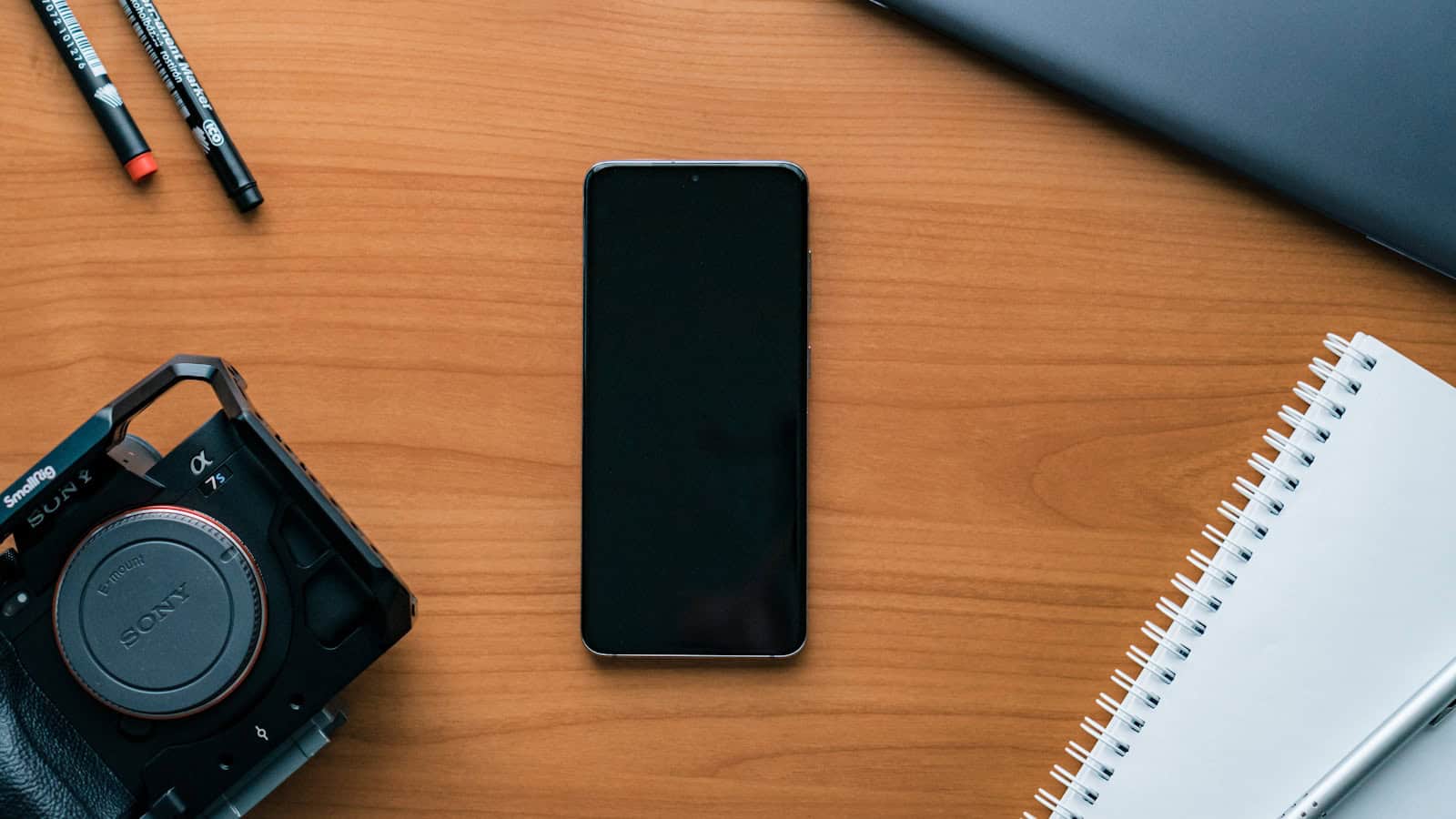 A dark phone lying face down on a wooden desk beside a notebook, pens and a camera.