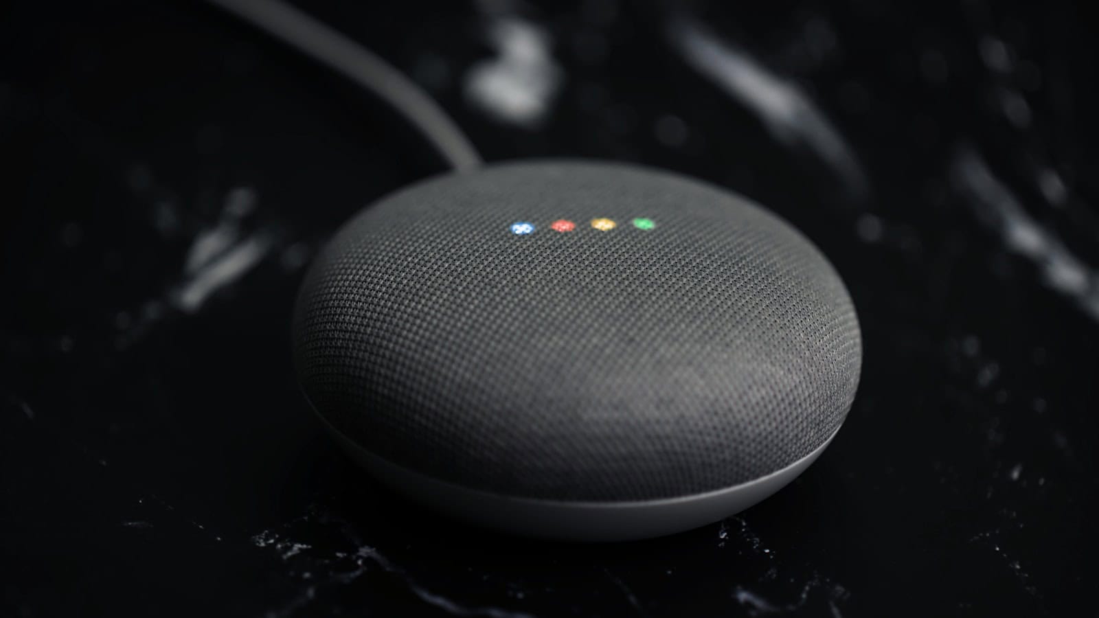 A dark fabric-covered smart speaker on a black surface with its four status lights lit.