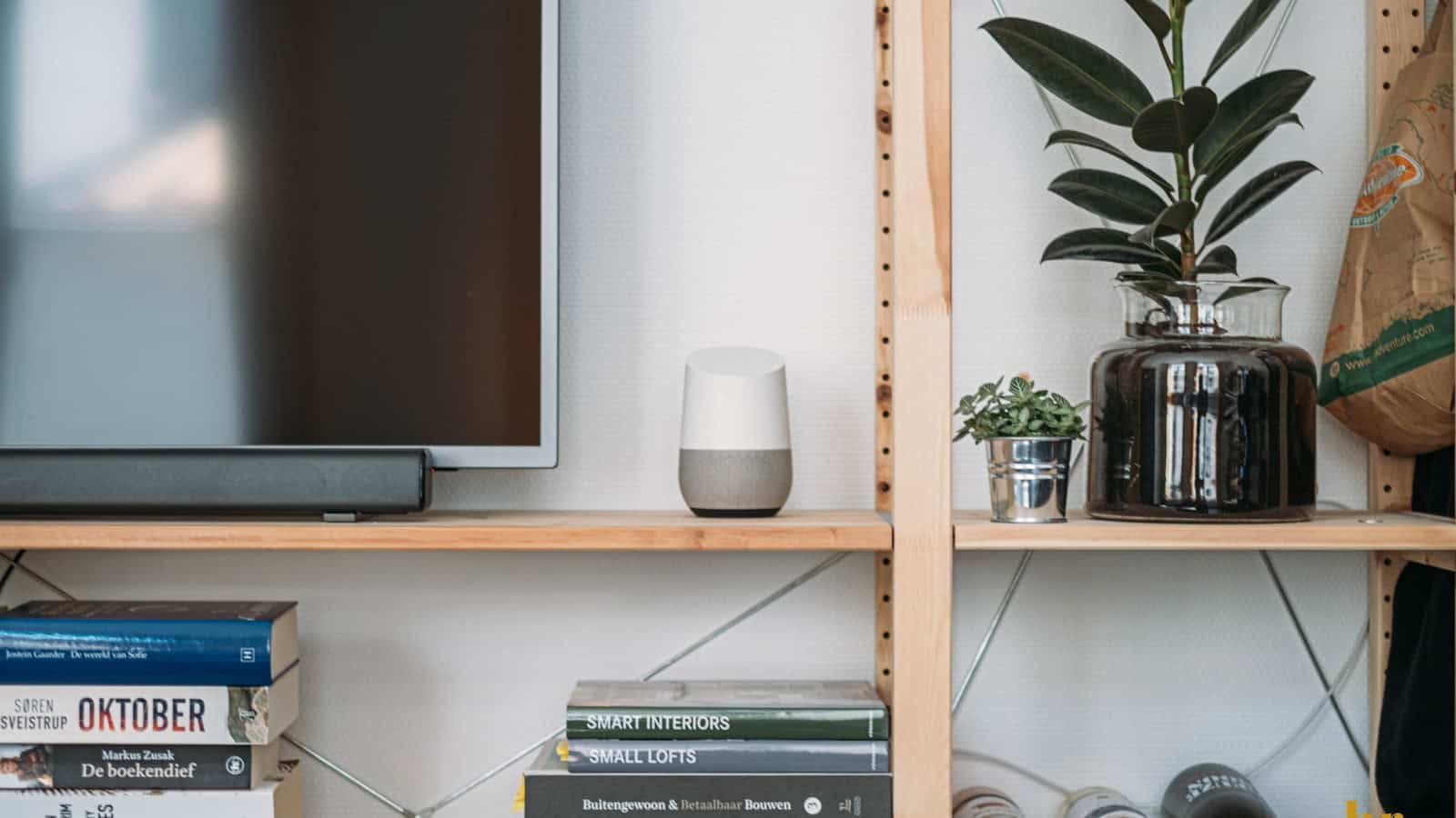 A white and grey smart speaker on a shelving unit beside a television and a row of books.