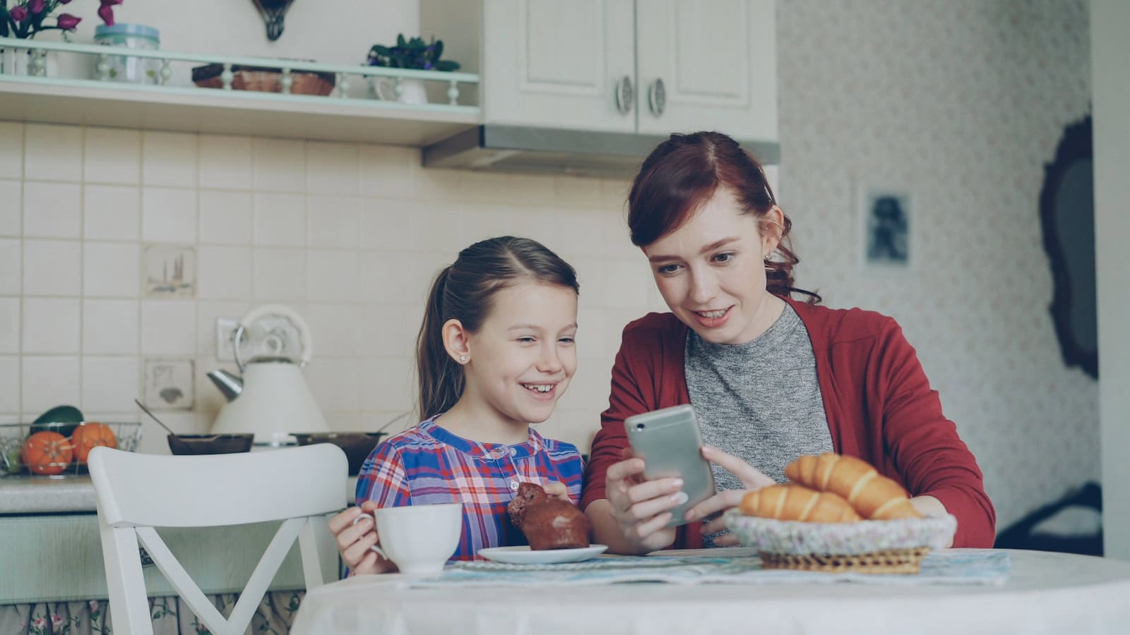 A mother and daughter at a kitchen table looking at a phone together.