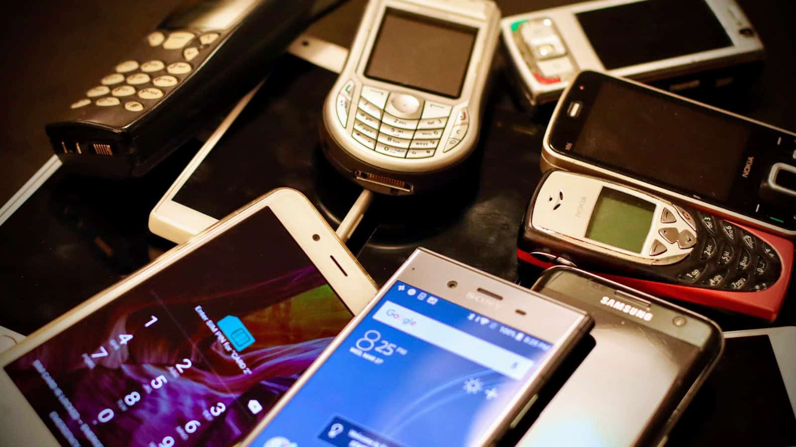 A heap of old and newer phones piled together, one of them still switched on.