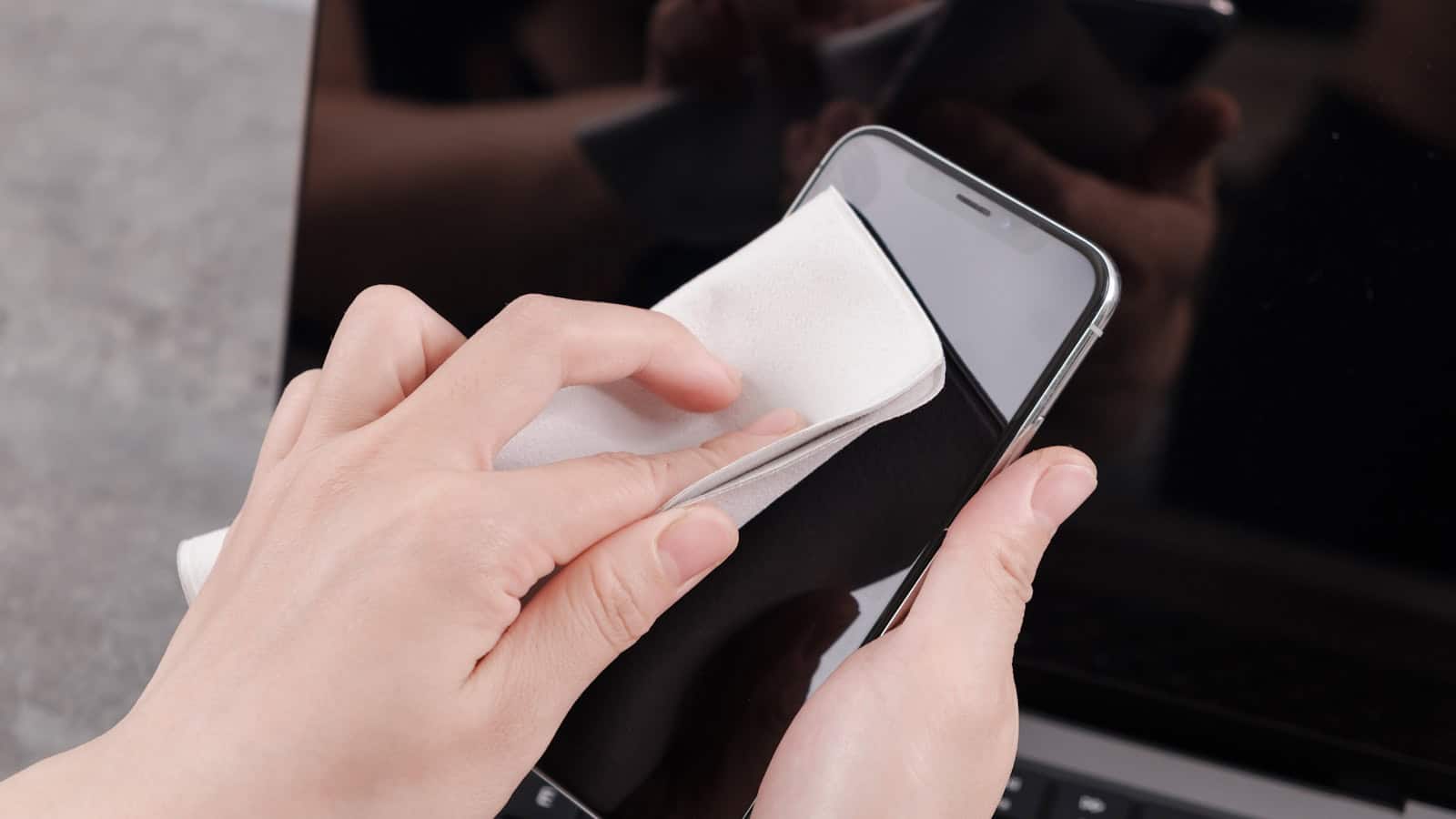 Two hands wiping the face of a dark phone with a folded white cloth.