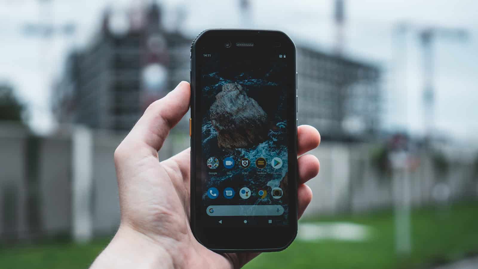 A rugged Android phone held up in one hand showing its home screen.