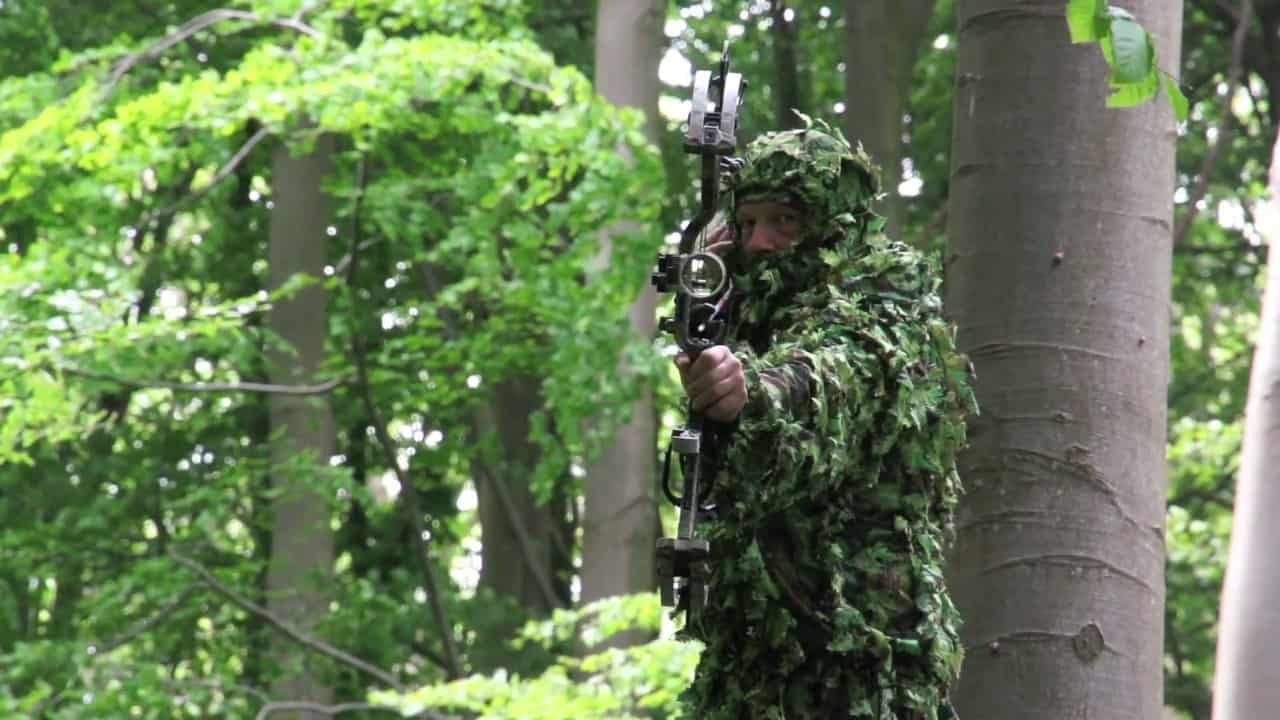 A camouflaged bowhunter drawing a compound bow in a green forest