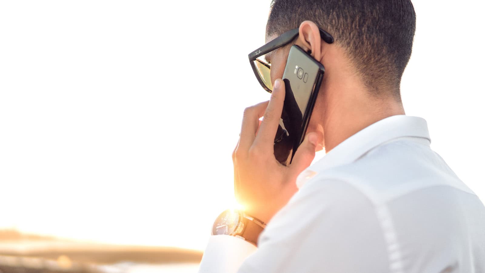 A man in a white shirt holding a phone to his ear outdoors at sunset.