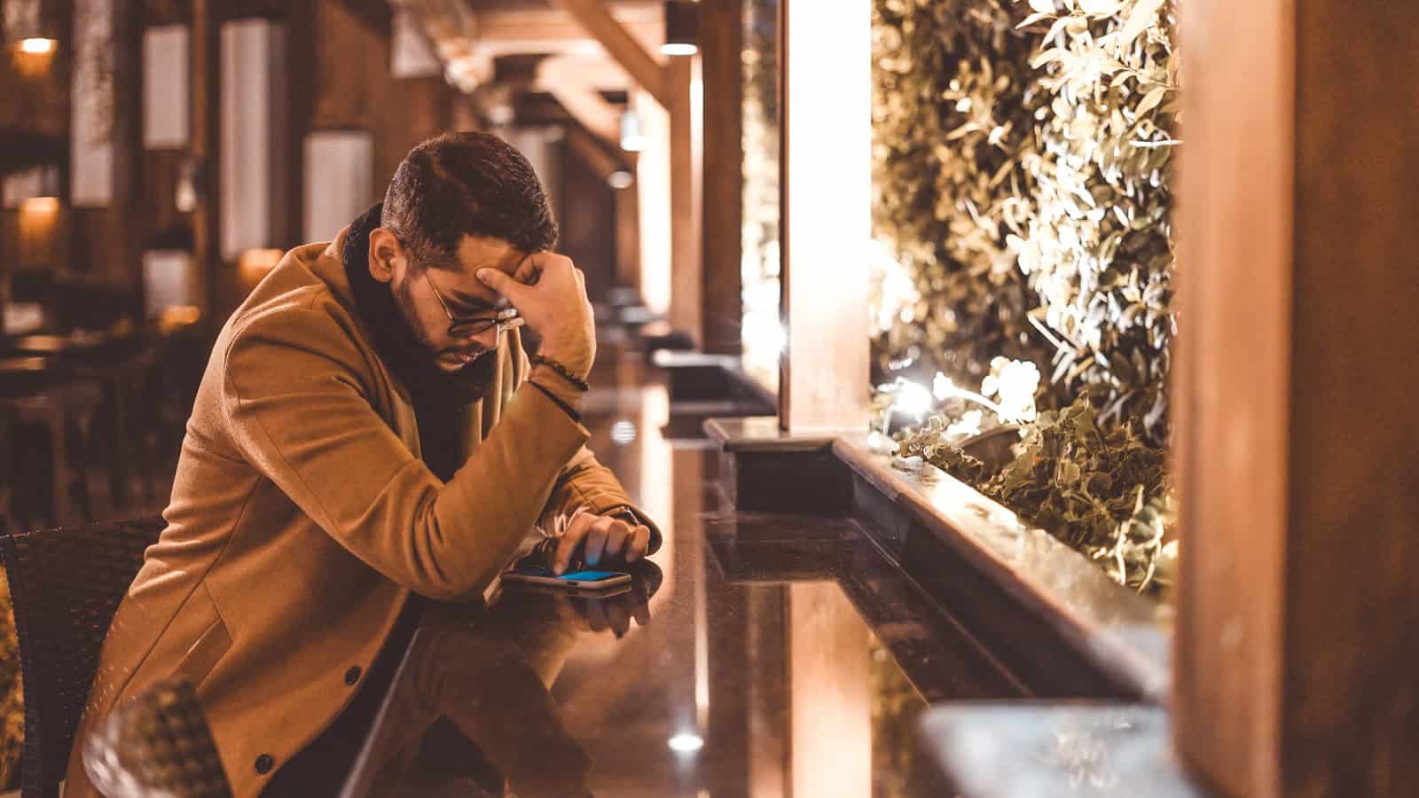 A man sitting at a counter with his head in his hand, looking down at his phone.