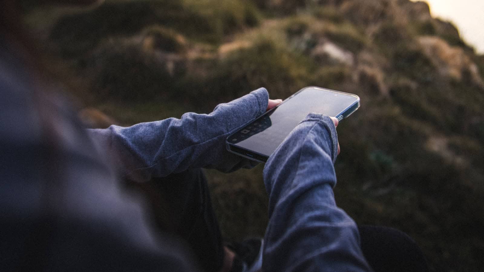 A person sitting on a hillside holding a phone with open moorland behind them.