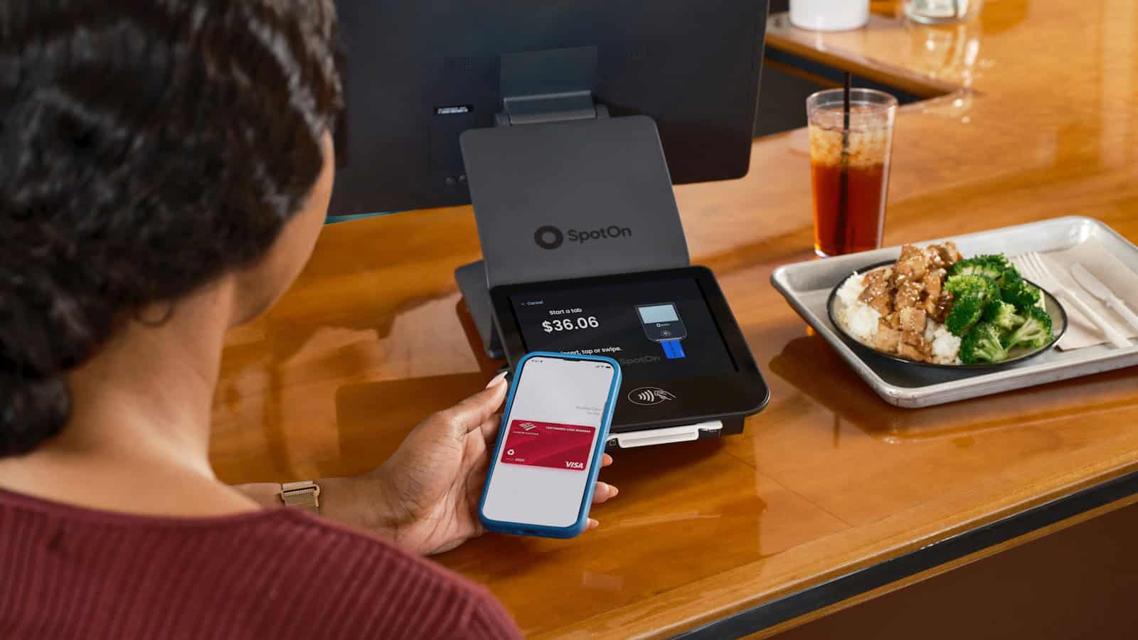 A woman holding a phone flat against a card terminal on a counter beside a plate of food.