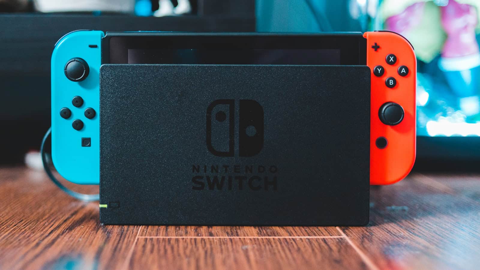 A Nintendo Switch seated in its dock on a wooden desk, Joy-Cons attached.