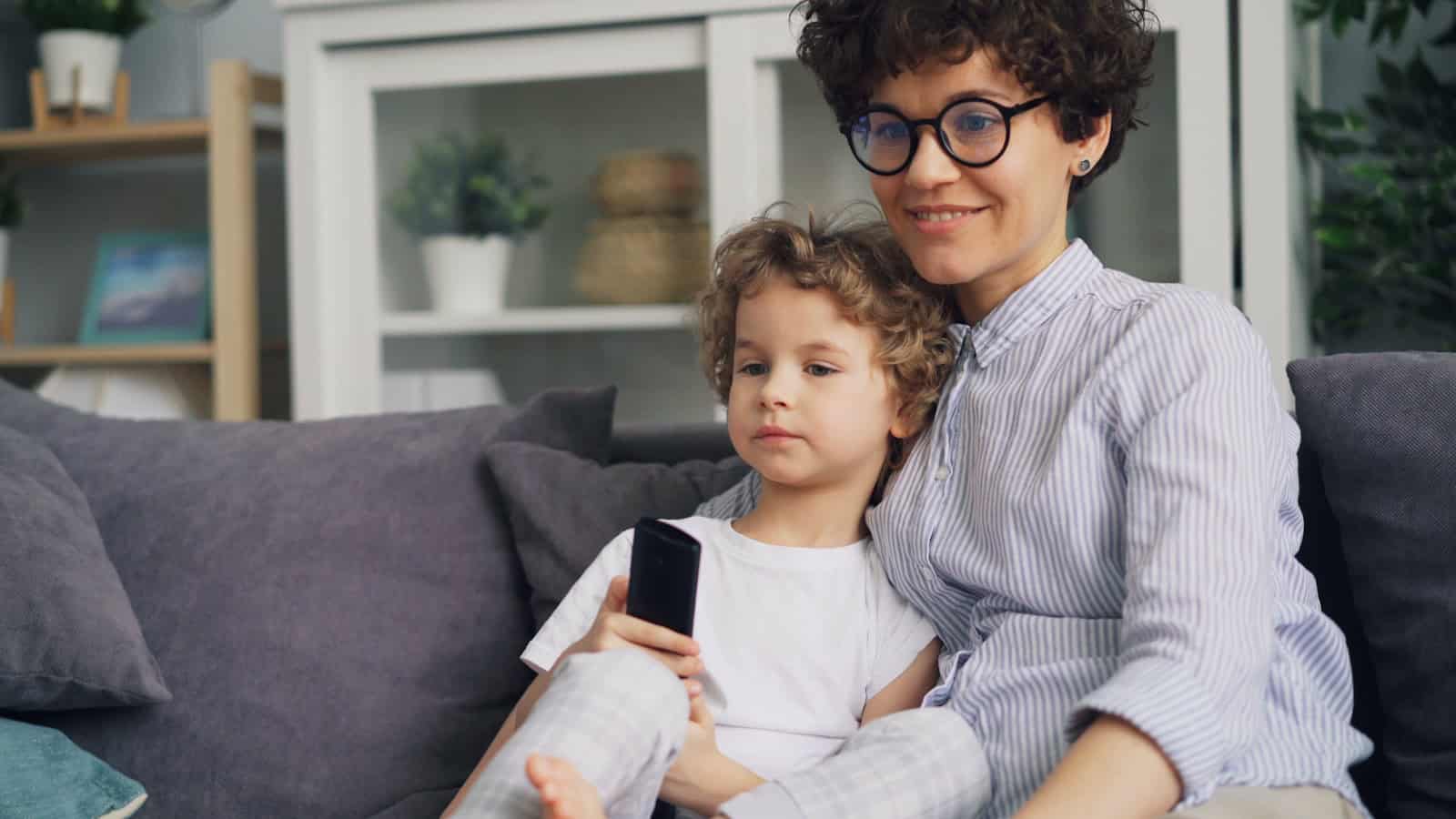 A parent sitting on a sofa with a small child on their lap, the child holding a phone.