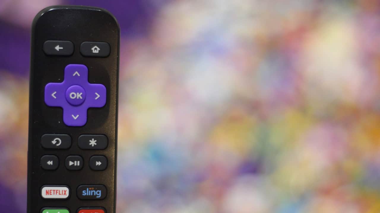 Close-up of a Roku remote showing the purple directional pad with OK in the centre, home and back buttons above, and app shortcut buttons below, against a blurred colourful background