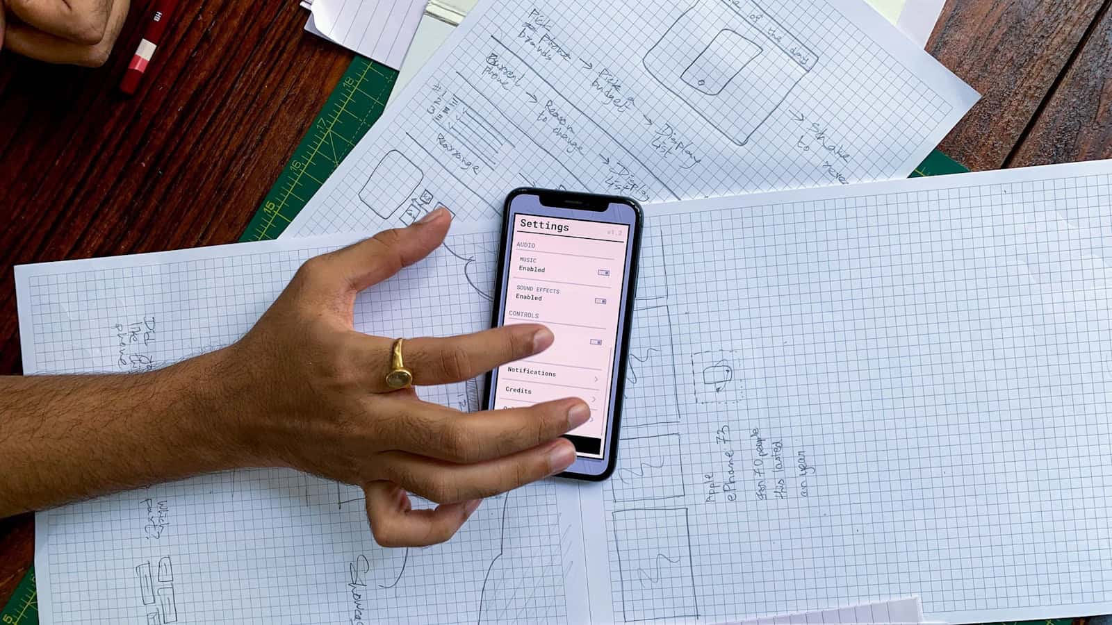 Two hands holding a phone over a desk covered in hand-drawn plans on graph paper.