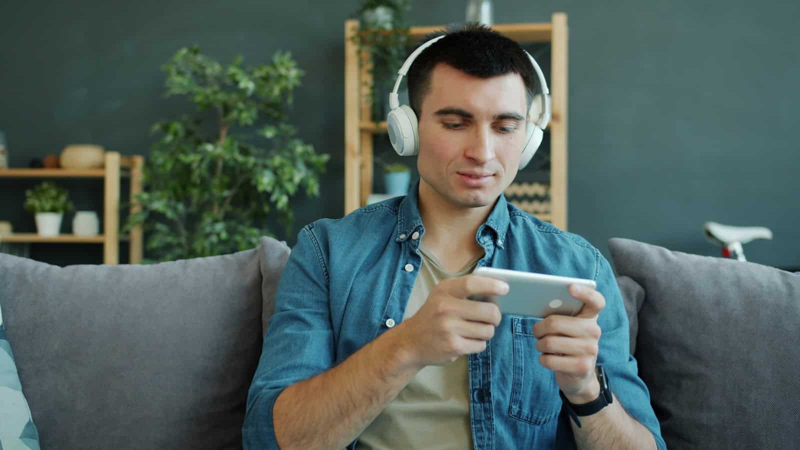 A man wearing over-ear headphones holding a phone sideways on a sofa.