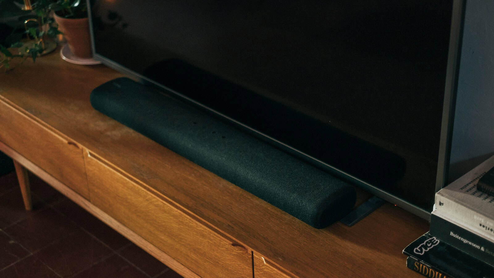 A black fabric-covered soundbar sitting on a wooden media console directly beneath a wall-mounted television that is switched off