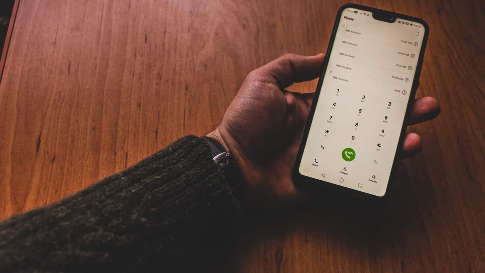 An Android phone held over a wooden table showing the dialer keypad above a list of recent calls.