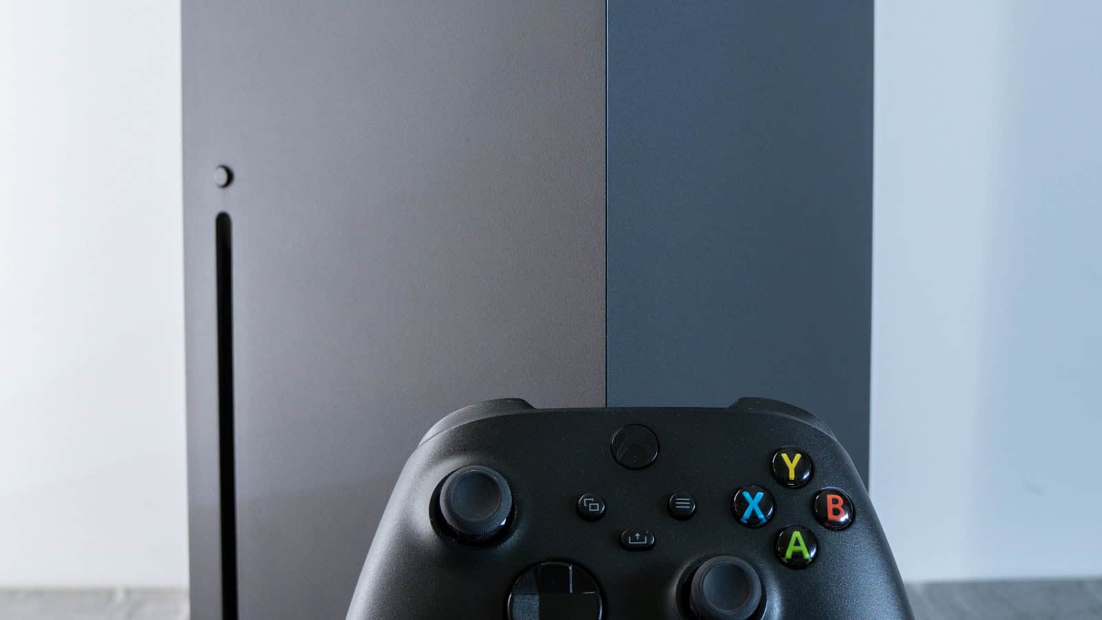 An Xbox Series X console standing beside a wireless controller against a plain wall.